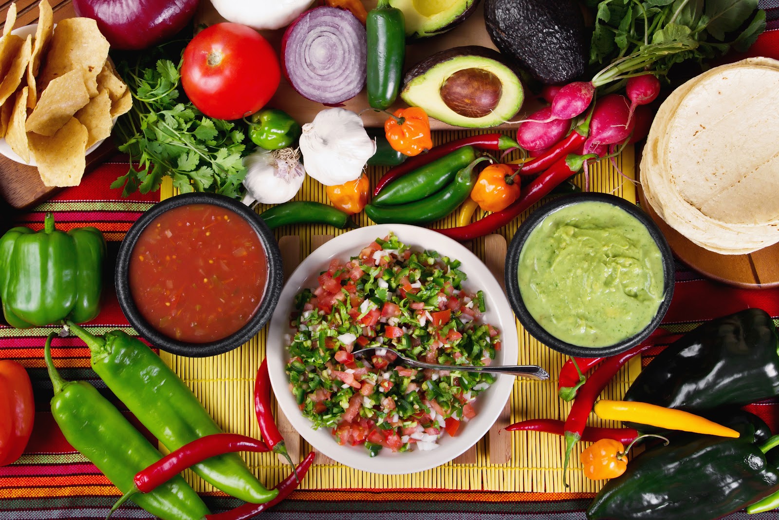 Fresh salsa, pico de gallo, and guacamole surrounded by peppers, tomatoes, and vegetables